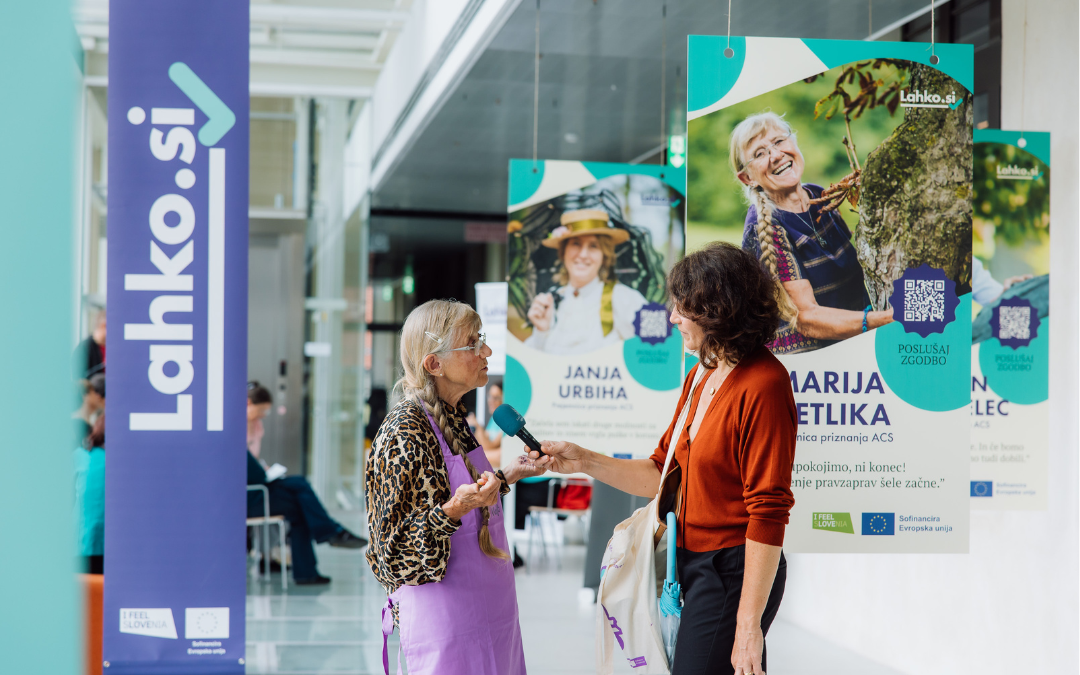 Na sliki se izvaja dogodek v notranjem prostoru, kjer poteka intervju. Na levi strani je vijoličen pano z napisom "Lahko.si", spodaj pa logotipa "I Feel Slovenia" in Evropske unije. Na sredini stojita starejša gospa oblečena v leopardji vzorec in vijolični predpasnik, ki odgovarja na vprašanja v mikrofon drugi mlajši gospe z rjavimi lasmi in oranžno jopico. V ozadju so razstavljeni veliki plakati z nasmejanimi obrazi žensk in informacijami, kot so imena, QR kode in kratka besedila.